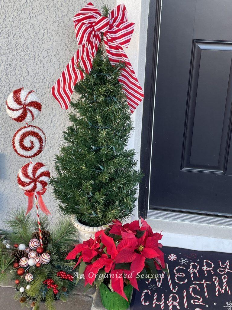A Christmas tree in an urn topped with a candy stripes ribbon and a poinsettia. An example of how to welcome guests with holiday porch decor.