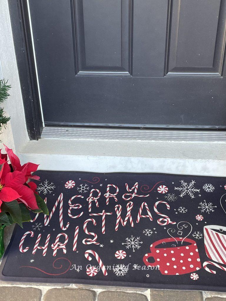 A black doormat that says Merry Christmas in candy cane letters. An example of how to welcome guests with a holiday porch decor.