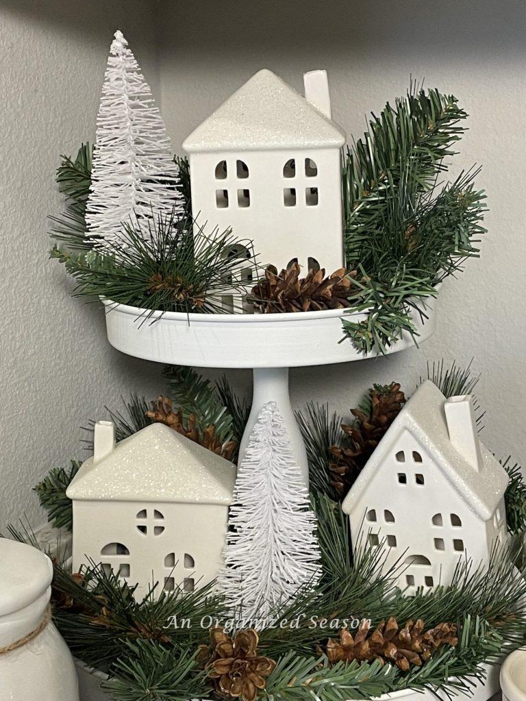 A tiered tray decorated with white houses, greenery, pinecones, and bottle brush trees adds a little bit of Christmas to the kitchen. 