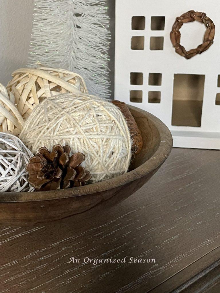 A bowl with yarn orbs and pinecones in front of a white house used to decorate a home for Winter. 