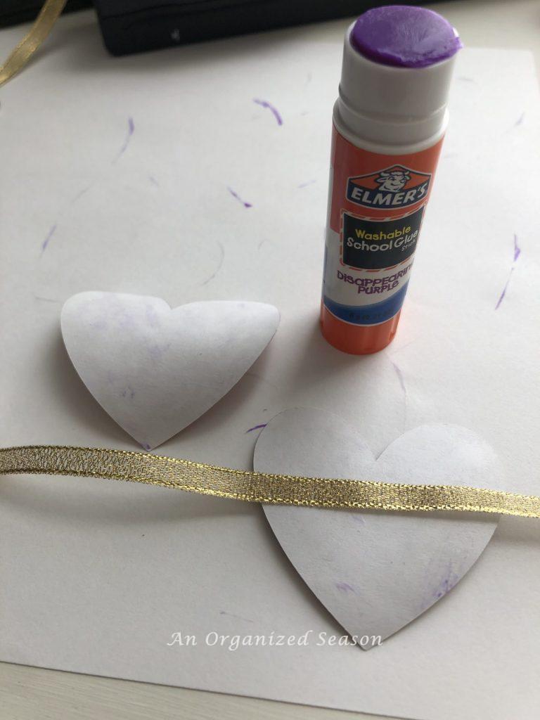 Two hearts, gold ribbon, and a glue stick used to make a paper heart and ribbon garland.