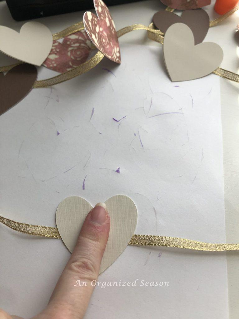 A woman's finger pressing two glued hearts onto a ribbon to make a paper heart and ribbon garland.