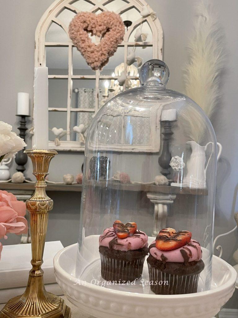 Table decor for Valentine's Day with a glass cloche covering chocolate cupcakes with pink icing and strawberry heart!