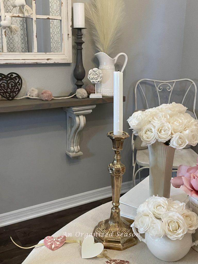A gold candlestick holder with white candle, next to vases of white roses on a table  displaying decor for Valentine's Day. 