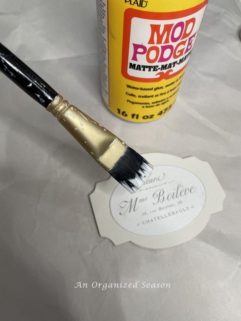 Applying Mod Podge to a wood tag, step five to make a floral basket wreath.