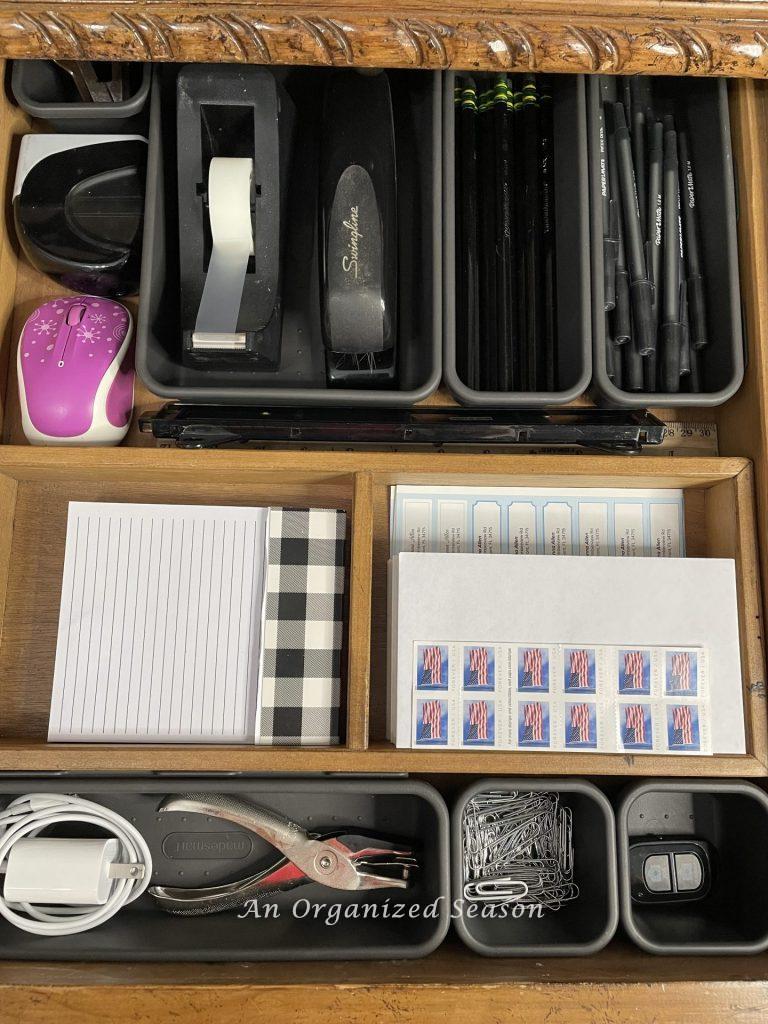 A desk drawer with black organizing bins used to separate supplies like pens, pencils, tape, paper clips. An example of ideas to organize a desk.