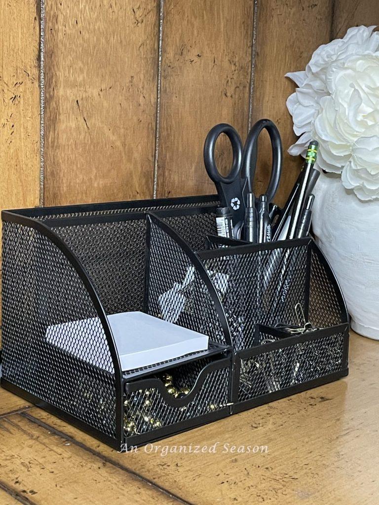 A black wire mesh desk organizer containing scissors, pens, paper clips, and post-it notes showing ideas to organize a desk.