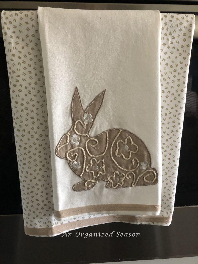 Two dish towels hanging on an oven door, to be used to make a no-sew dish towel pillow. One has a bunny and the other has bunny tracks. 