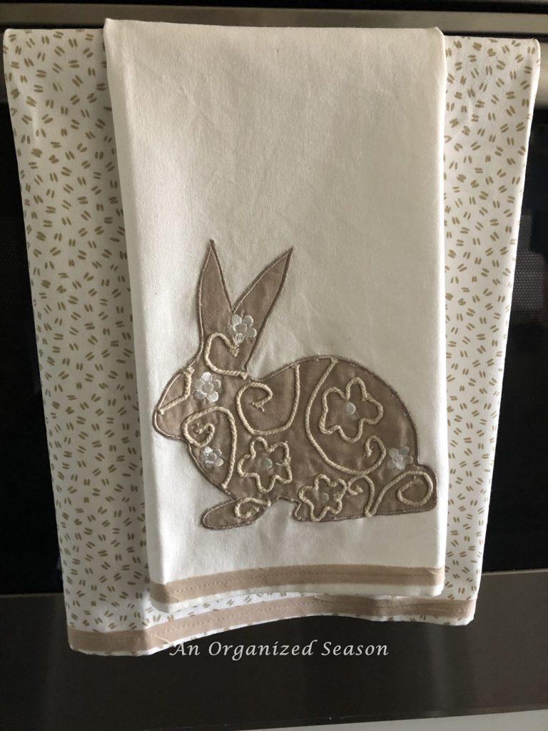 Two dish towels hanging on an oven door, to be used to make a no-sew dish towel pillow. One has a bunny and the other has bunny tracks.