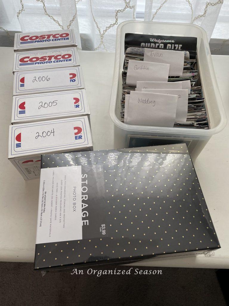 Five Costco photo boxes and a plastic bin full of photographs sitting next to a new photo box, showing ideas to organize a craft space.