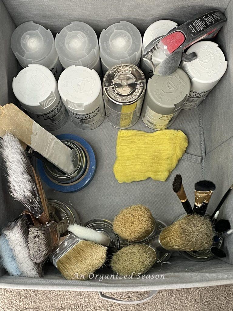 A fabric cube holding spray paint cans and jars of paint brushes, showing ideas to organize a craft space.