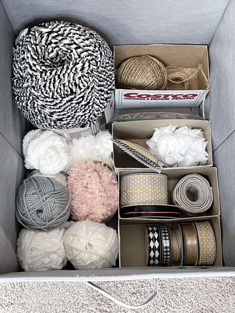 A fabric cube bin holding yarn and four Costco photo boxes used to separate ribbons, an example of ideas to organize a craft space.