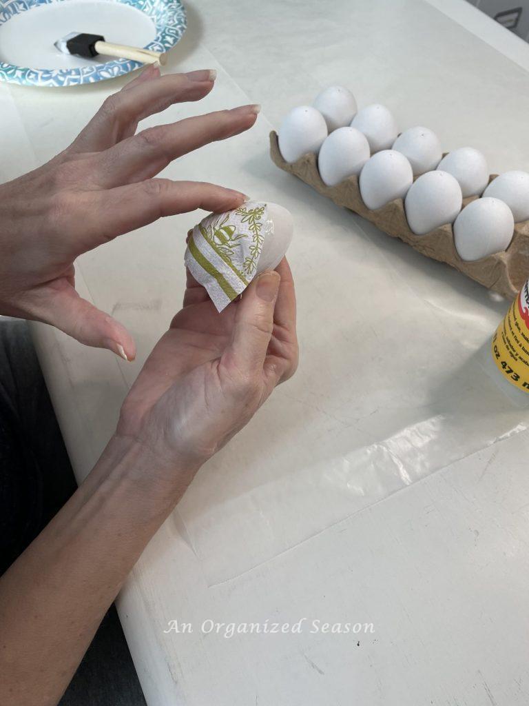 A woman using her finger to adhere a napkin piece onto an egg showing how to do a  decoupage Easter egg DIY.