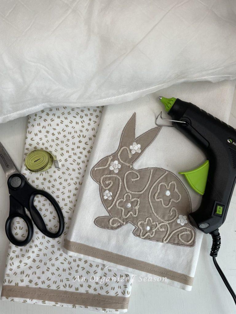 Two dish towels, a pillow insert, hot glue gun, scissors, and a measuring tape, used to make a no-sew dish towel pillow. 