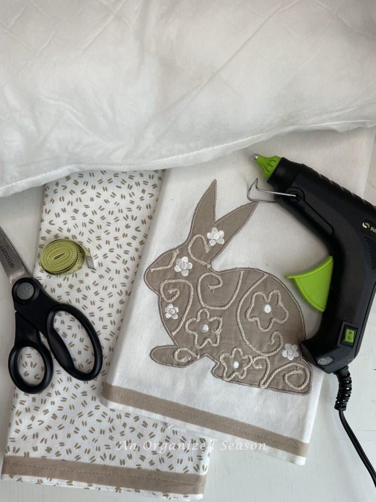 Two dish towels, a pillow insert, hot glue gun, scissors, and a measuring tape, used to make a no-sew dish towel pillow.