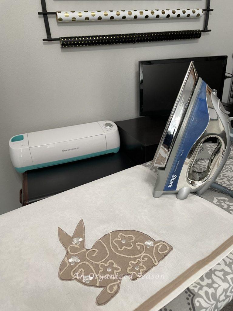 A dish towel on an ironing board with an iron next to it, that will be used to make a no-sew dish towel pillow.