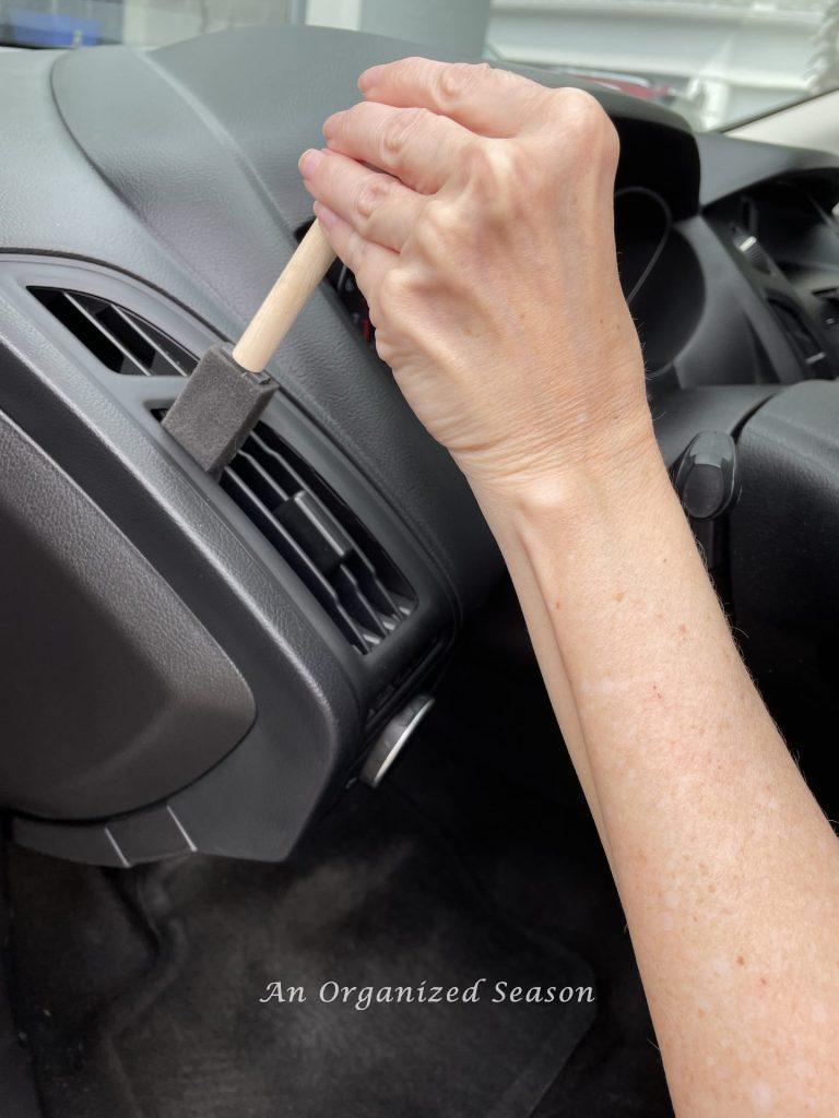 Someone using a small foam paintbrush to clean the vents of a car, showing one strategy to clean and organize a car.