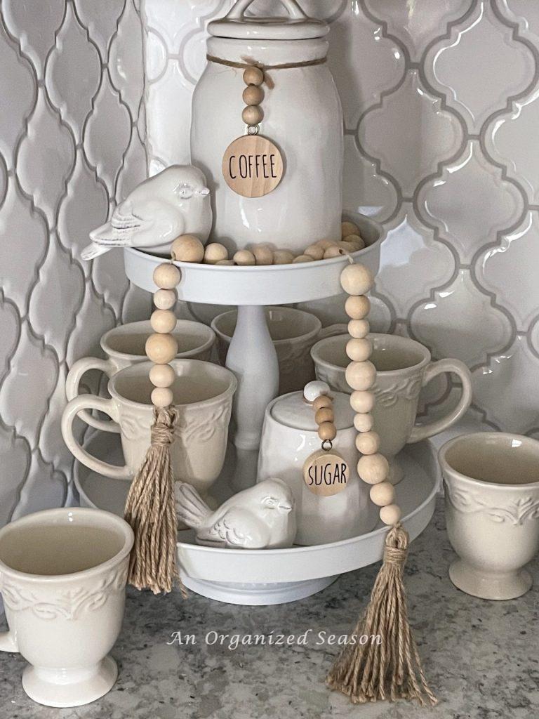 A white tiered tray with tea canisters, coffee cups, two ceramic birds, and a wood bead garland showing ideas to decorate your home for Spring.