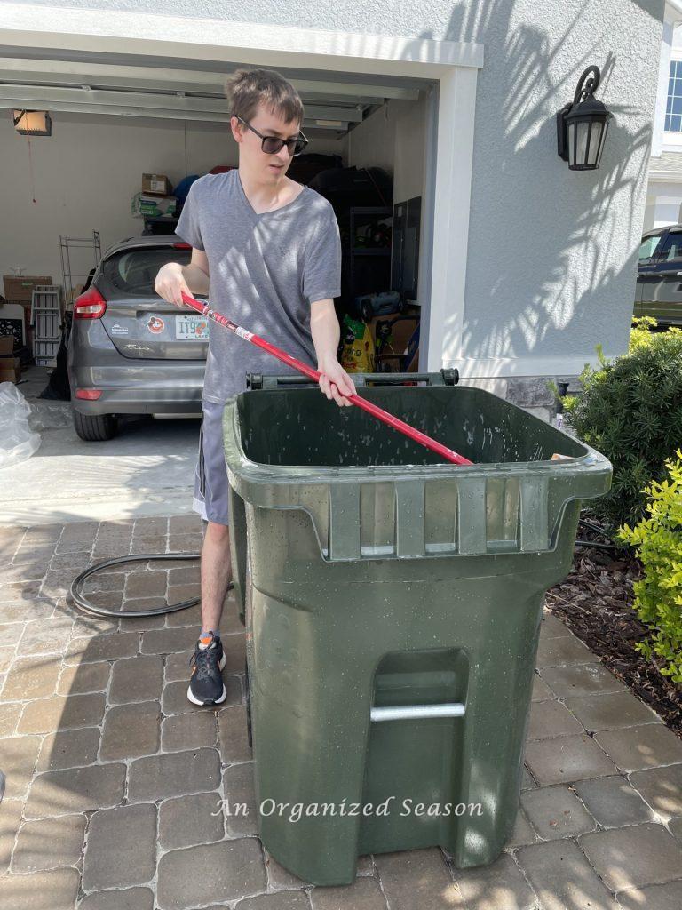 A man cleaning a trash can, outside a garage, with a broom. Showing how to organize a garage into zones.