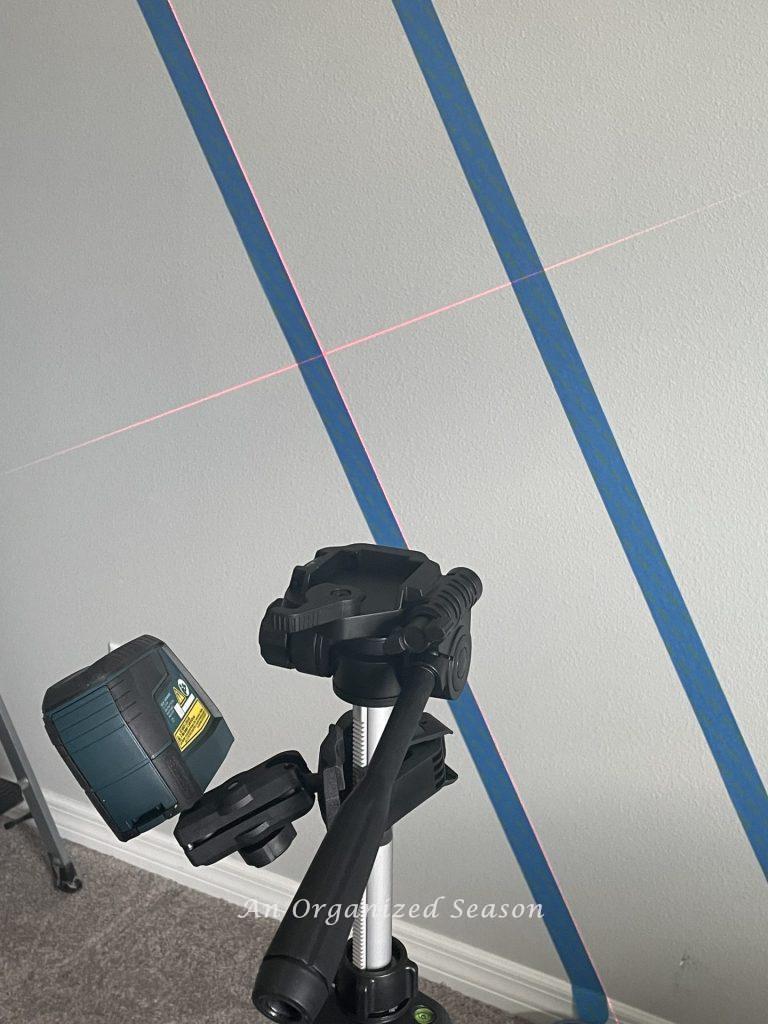 A laser level mounted on a tripod shining light on a wall to build a modern accent wall in a bedroom. 