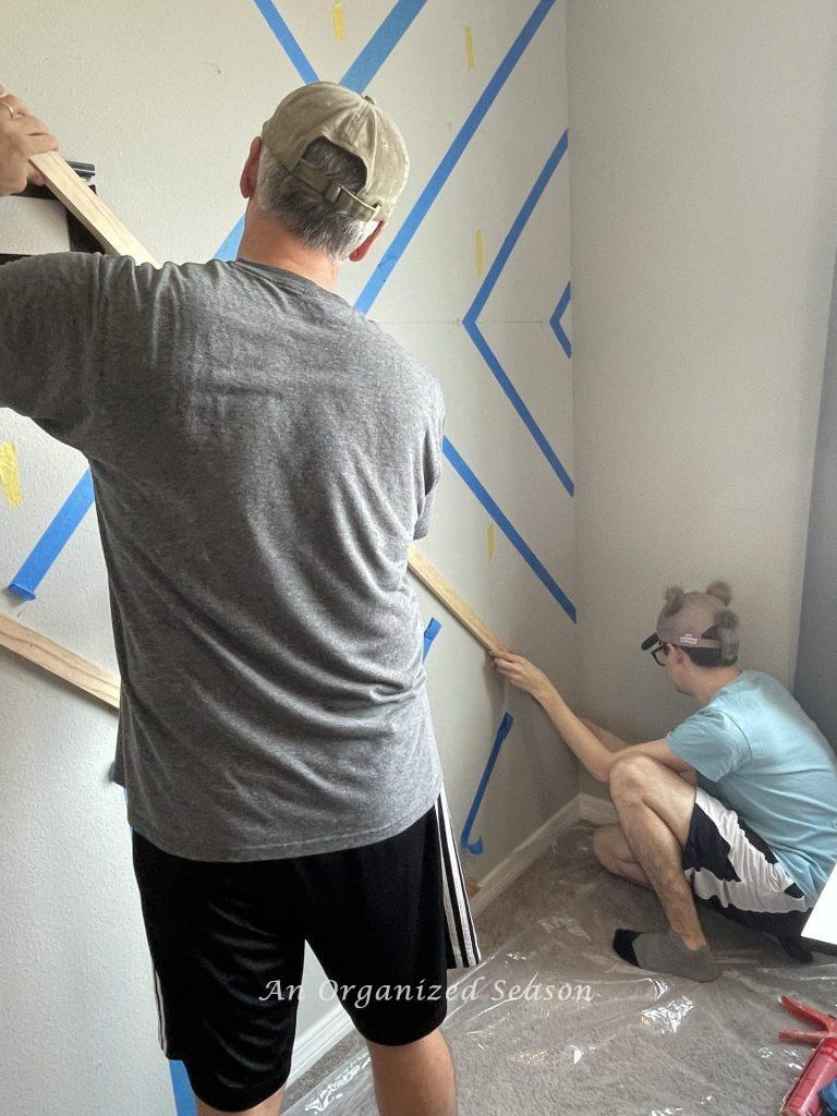 Two men replacing painters tape with 2 inch boards, step two to build a modern accent wall in a bedroom.