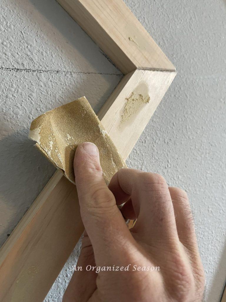 A man sanding wood filler on a board, step five to build a modern accent wall in a bedroom.