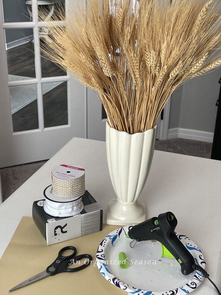 Dried wheat in a vase, hot glue gun, ribbon, and scissors used to make an arrangement. 