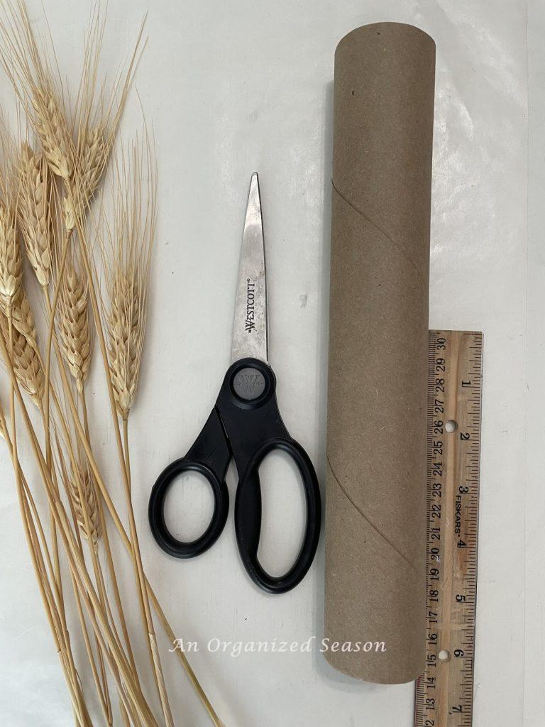 An empty paper towel roll, scissors, a ruler, and dried wheat used to make an arrangement.