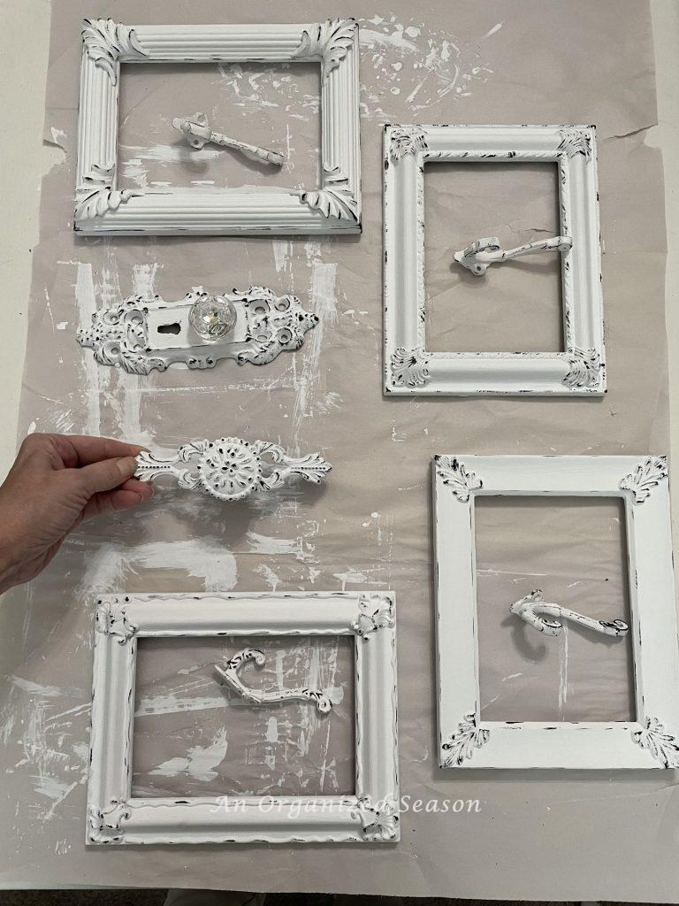 Four white frames, four hooks, a door knob, and cabinet pull used to make DIY towel hooks. 
