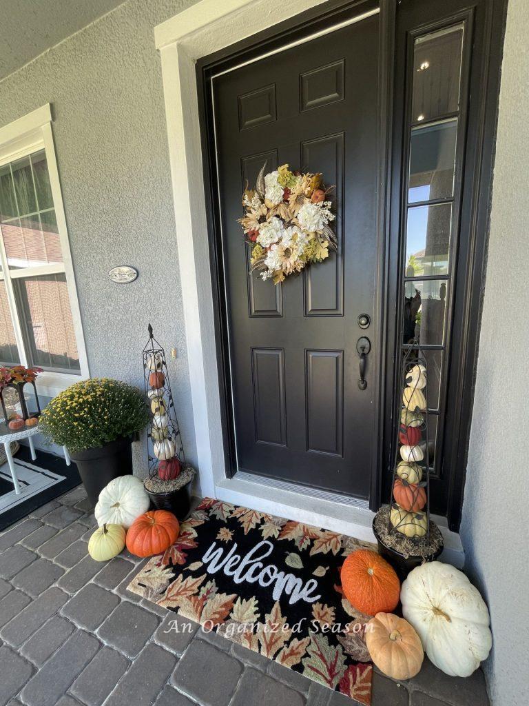 A porch decorated with two pumpkin obelisks, assorted pumpkins, a mum. a welcome rug, and a wreath. 