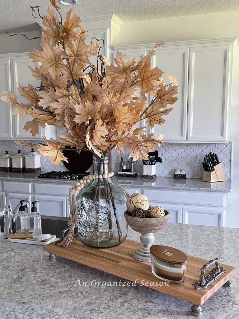 Glass vase filled with maple leaves, sitting on a wood tray in a kitchen.