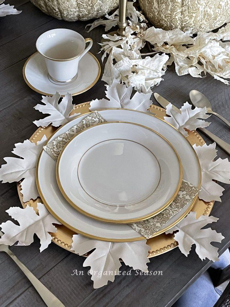 A table setting with cream colored china on a gold charger with white leaves. 