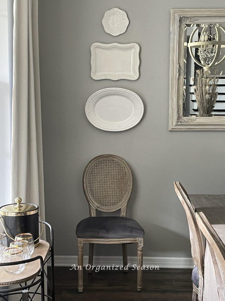 Three white plates hanging on a dining room wall above a Louis the 14th chair. 