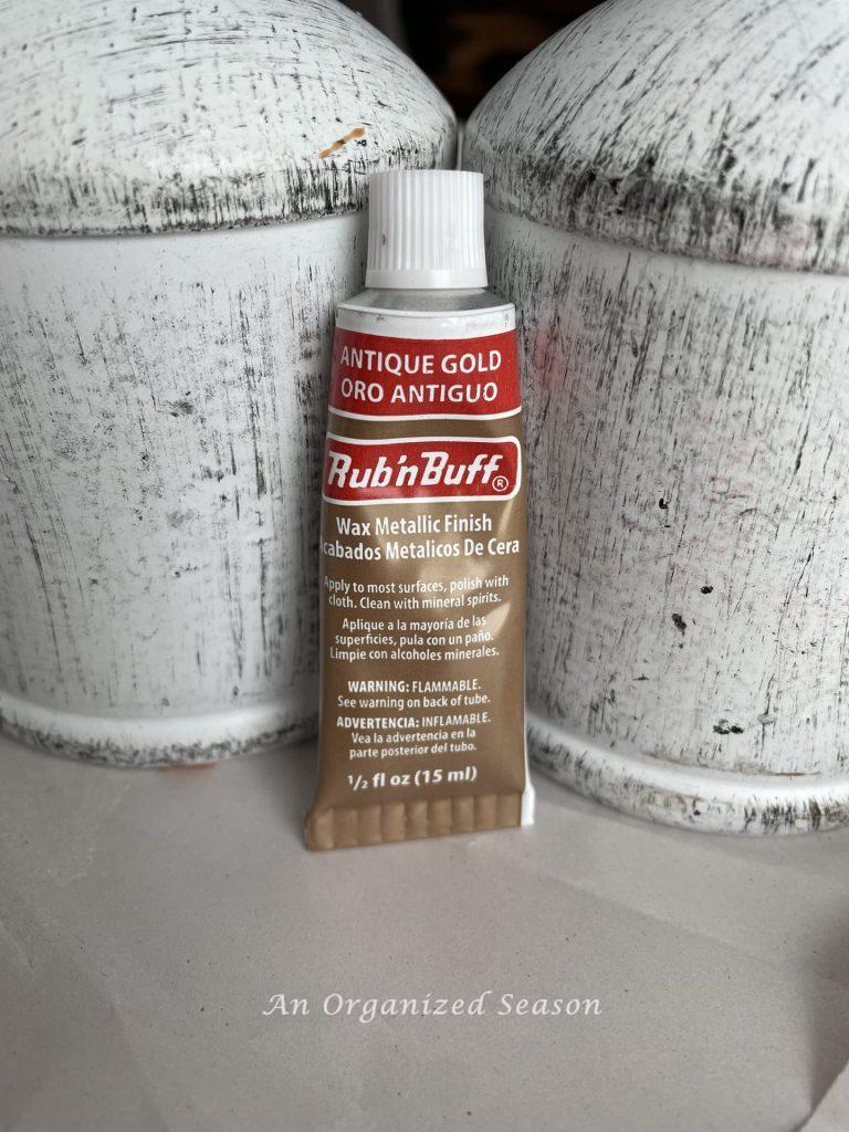 A tube of Rub 'n Buff sitting in front of white bells used to create a Pottery Barn rustic iron bell hack.