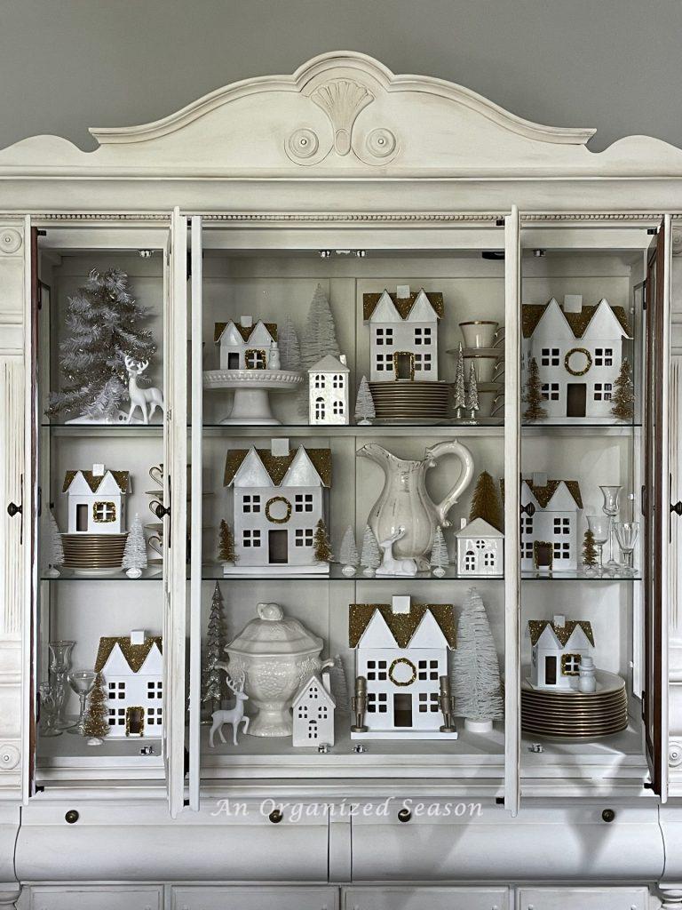A white and gold Christmas village styled in a white china cabinet. 