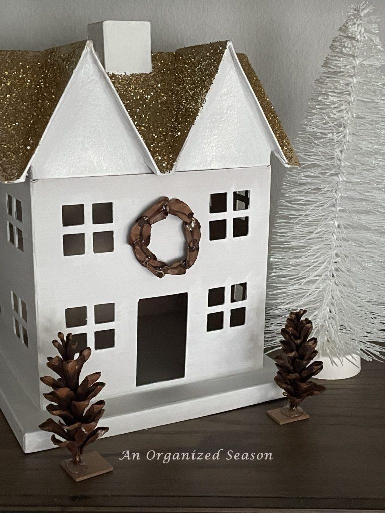 White house in a Christmas village embellished with a pine cone wreath and trees. 