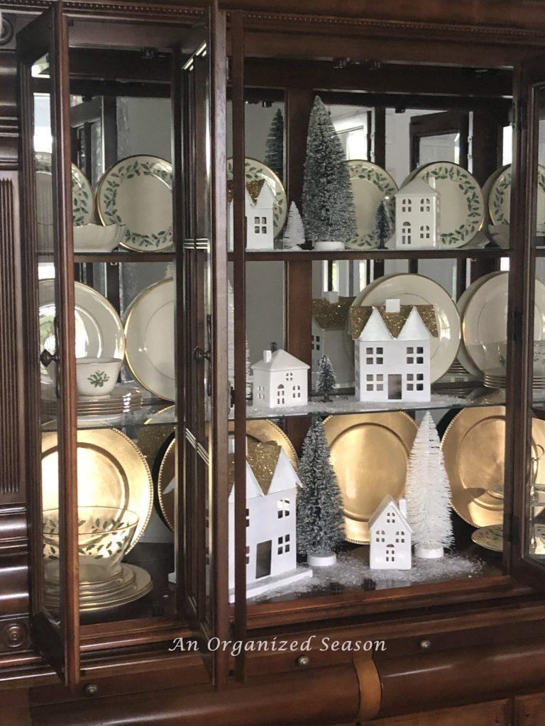 A small white Christmas village styled in a brown china cabinet.