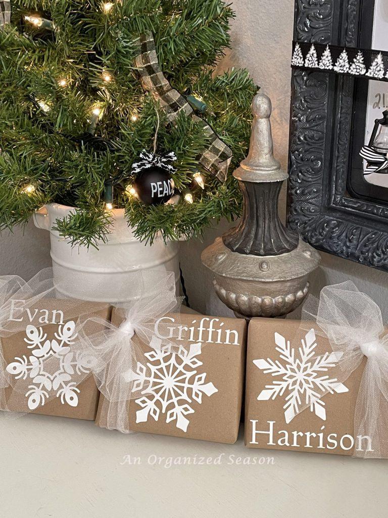 Three Christmas gifts wrapped in brown  craft paper embellished with white snowflake stickers and names. 