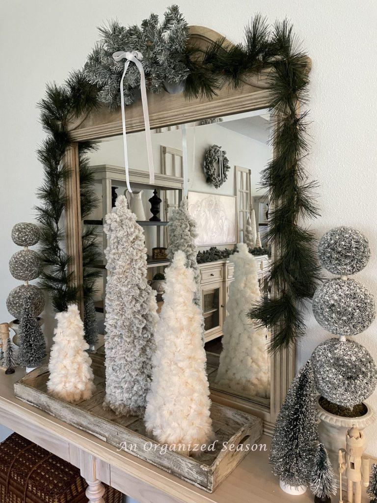 A console table and mirror decorated with evergreen garland, yarn cone trees, topiaries, bottle brush trees, and nutcrackers! 