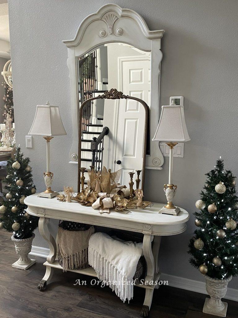 A table and mirror in a foyer decorated for Christmas, with  a small tree on either side of it. 