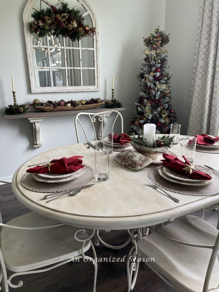 An eat-in kitchen with a tree, swag on a mirror, dough bowl on a shelf, and a table set with white dishes and burgundy napkins. 