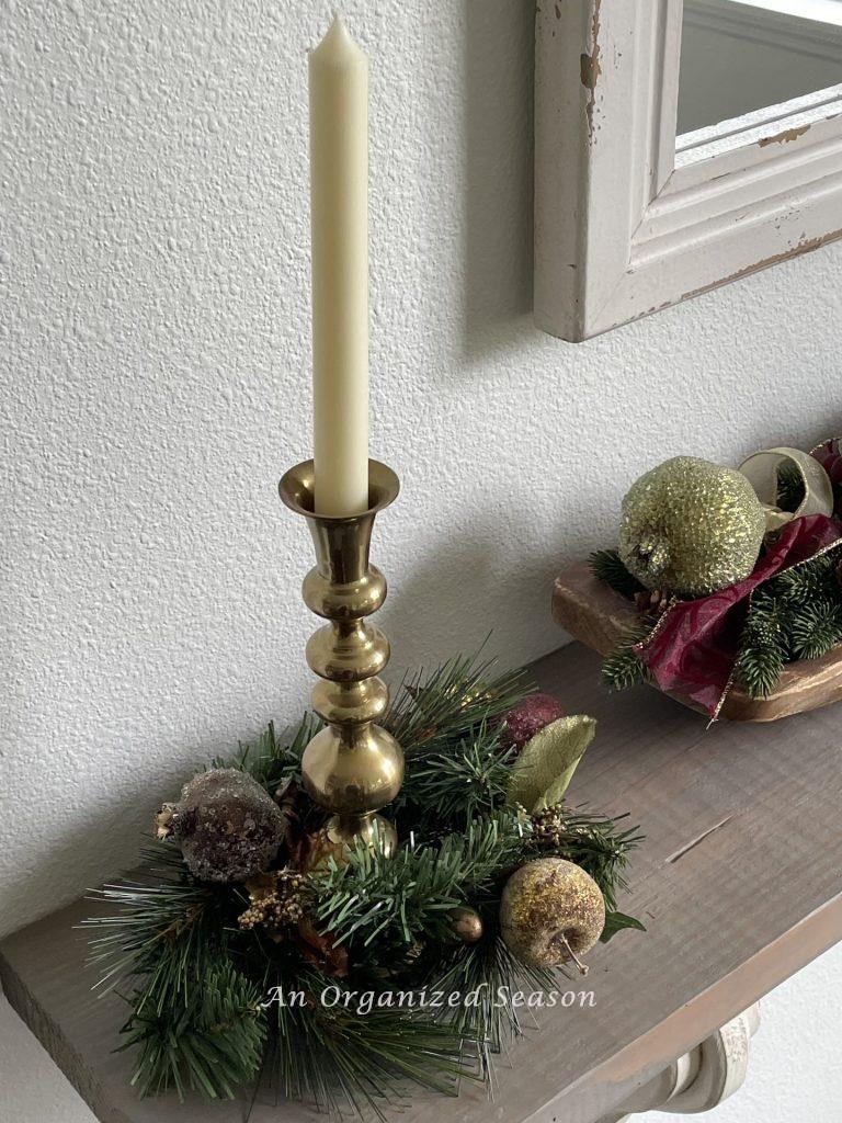 A taper candle in a brass candlestick surrounded by evergreen and faux fruit. 