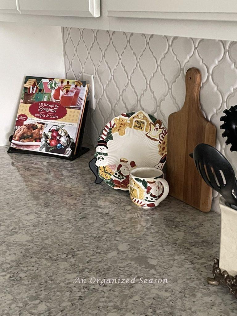 A Christmas cookbook on a stand and a plate and mug for cookies and milk for Santa, sit on a kitchen counter. 