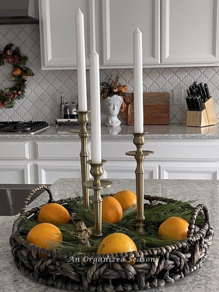 A round basket with three gold candlesticks, evergreen, and oranges decorate a kitchen counter for Christmas. 