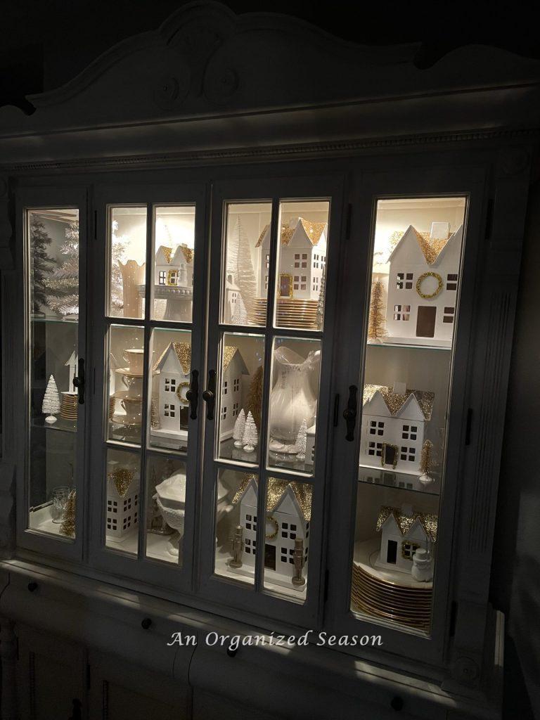 A China cabinet filled with a white Christmas village and lit up during the  nighttime home tour.