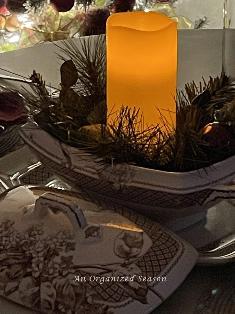 A lit pillar candle with evergreen and small faux fruit inside a brown transferware bowl, sitting on a table. 