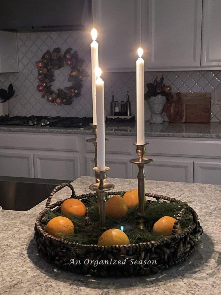 A round basket holds evergreen pieces, three brass candle holders with lit candles, and oranges on a kitchen counter.  