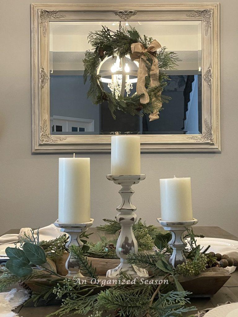 A wreath hanging on a dining room mirror with three candles on the table.