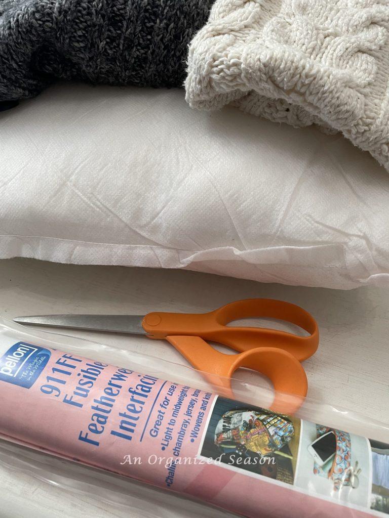 Sweaters, a pillow insert, scissors, and interfacing that will be used to make cozy home decor with sweaters.