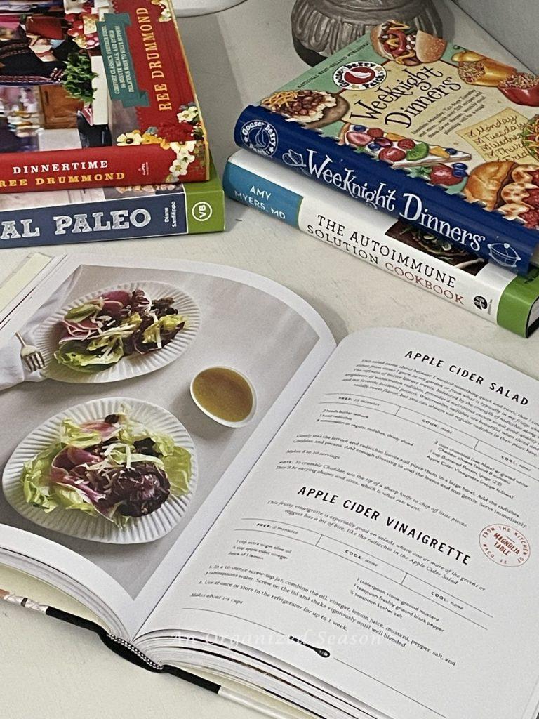 Five cookbooks on a table. One is open to a salad recipe. 